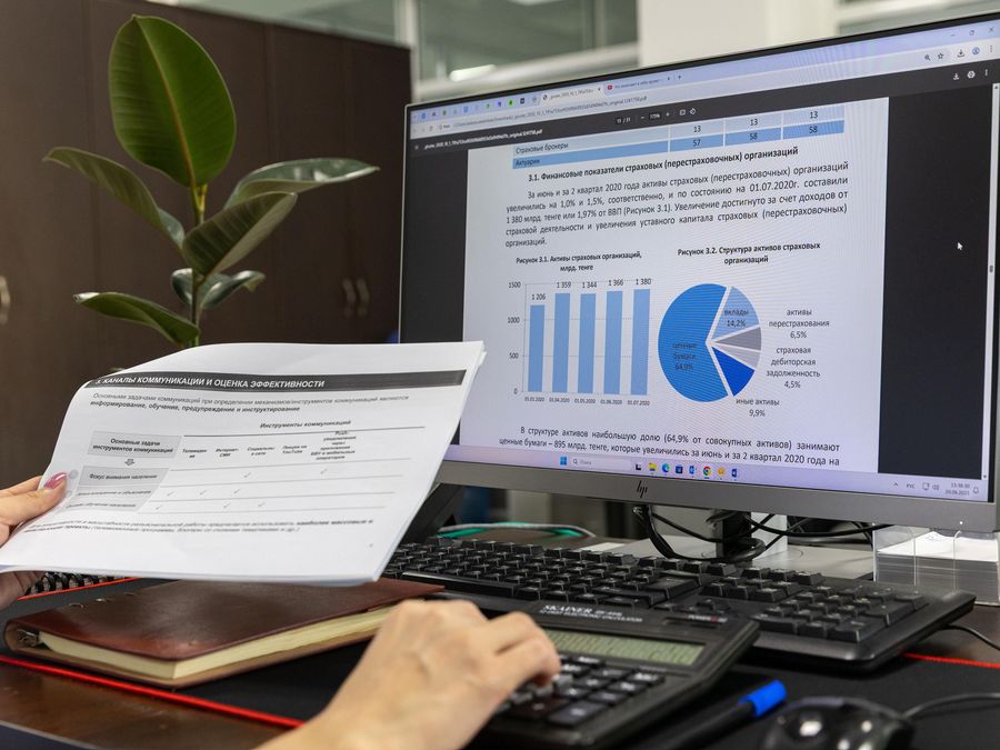 Unas manos sostienen un informe impreso junto a un monitor con tablas y gráficos.